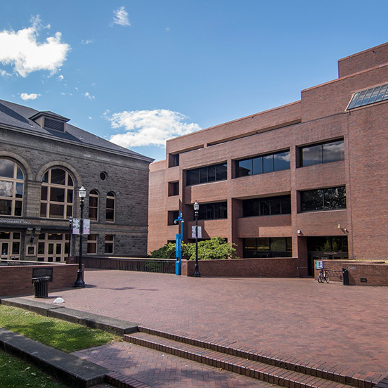  A large reddish-brown brick building stands next to a light-gray stone building at a right angle. In front of them, a brick-paved space extends.