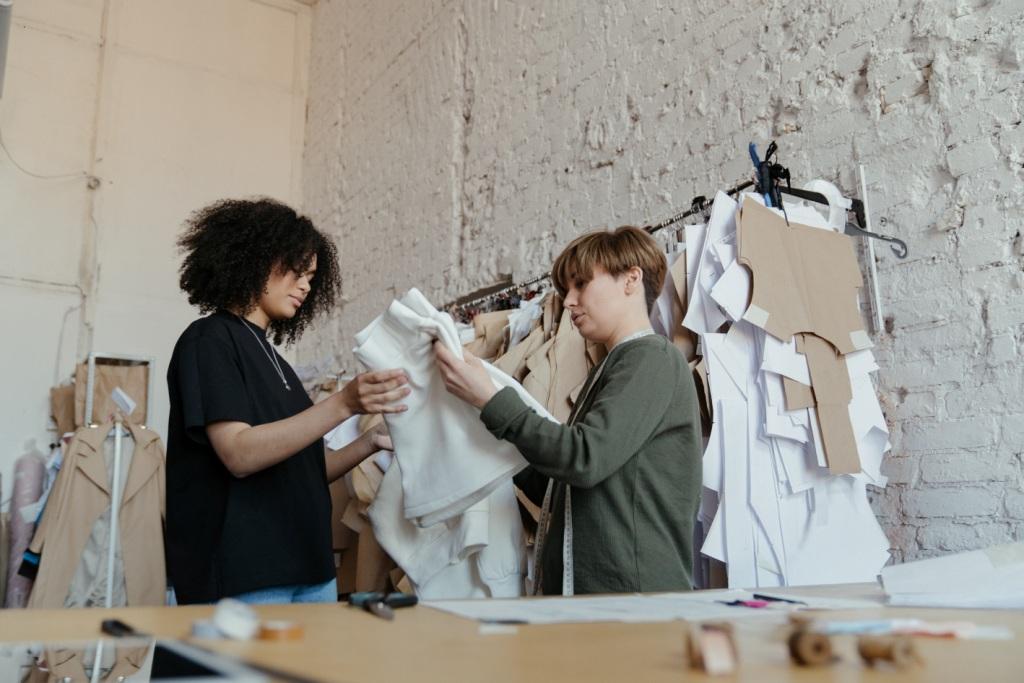 Two adults in a clothing design studio examine white fabric together at a worktable. Sewing materials and garments hang on the walls behind them.