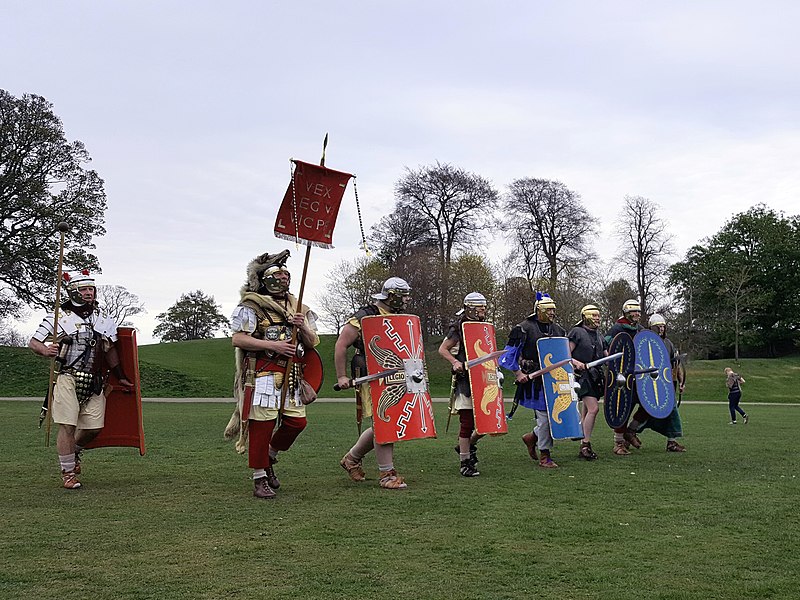 Line of six people dressed as ancient Roman army walking across grassy field 
