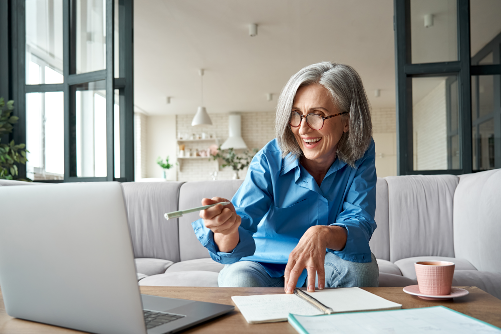 Photo of smiling woman sitting on couch with notebook on table using pencil to point at open laptop 