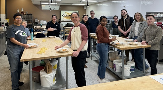 Several students standing around two long tables in an industrial kitchen, smiling at camera. 
