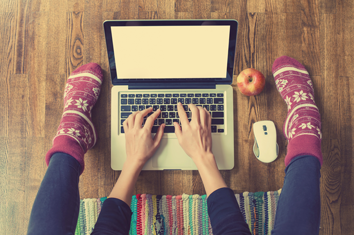 Laptop computer has hands on keyboard and feet/leggs on either side, wooden floor below. 