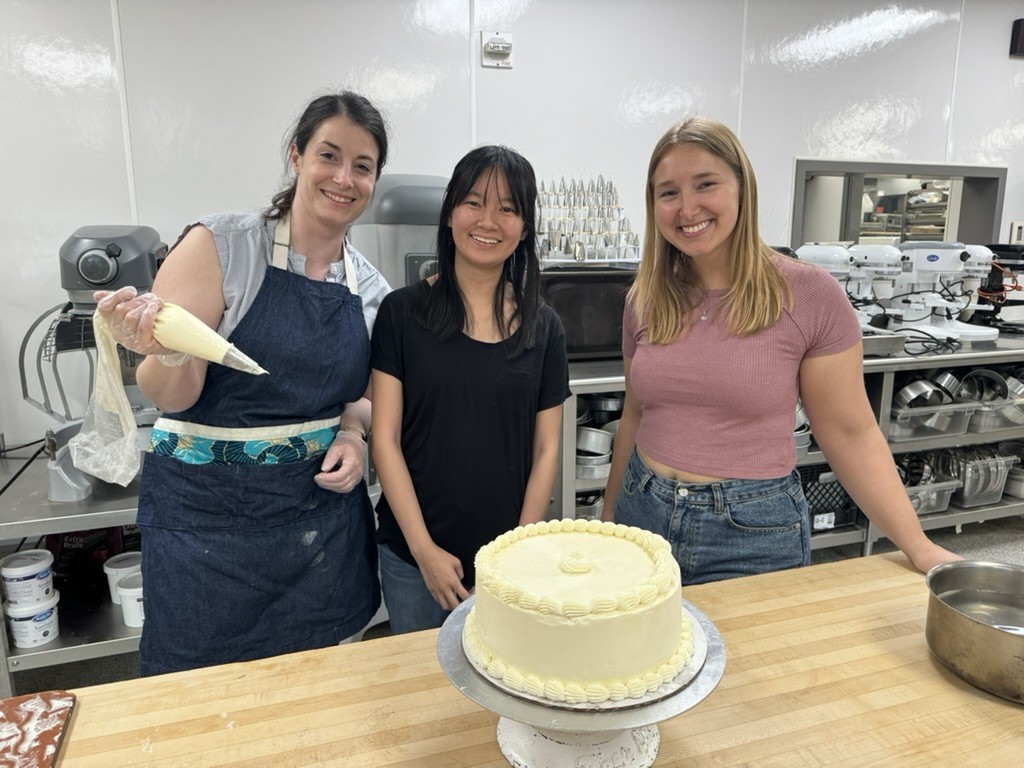 Three women in a kitchen stand at wooden counter with white frosted cake on platter. 