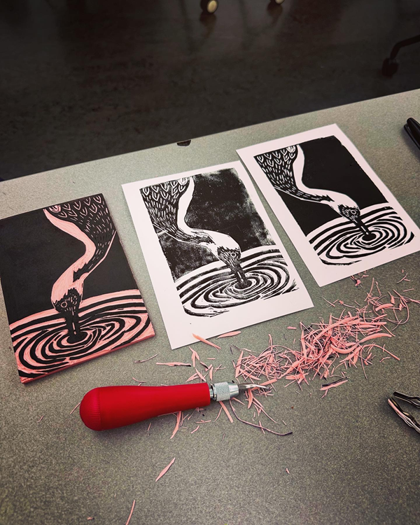 Pink linoleum block with ink sits next to to the image printed on paper (two) with a red carving tool in the foreground.