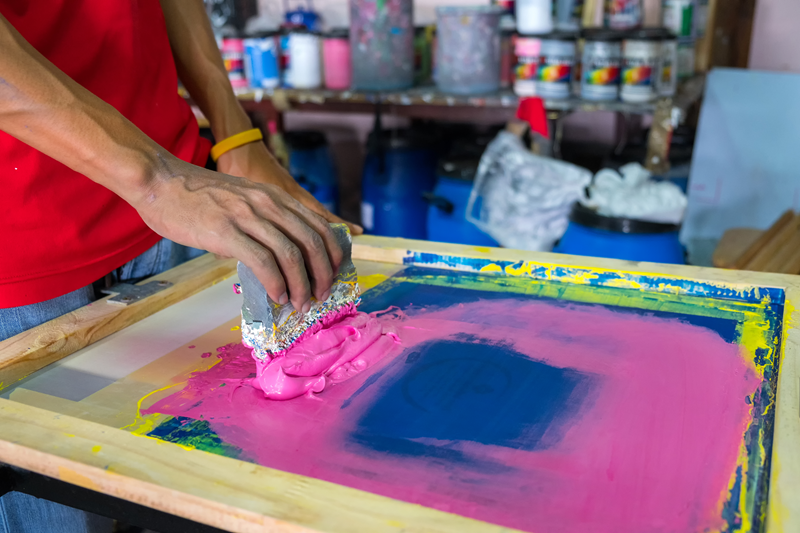 Person on left has both hands on scraper tool that sits on screen printing screen with pink ink and blue in background. 