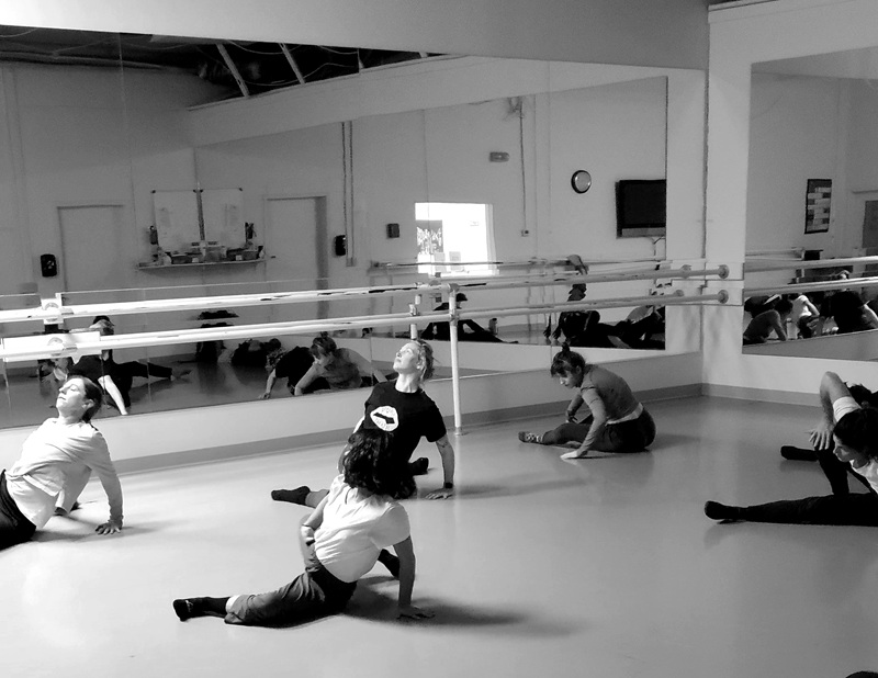 People stretching on floor with one leg behind and one in front, facing upwards, black and white, in dance studio. 