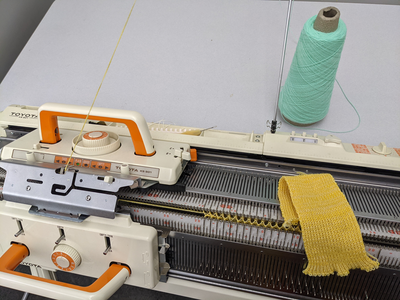 Knitting machine with bobbin of light teal thread and piece of yellow knit fabric. 