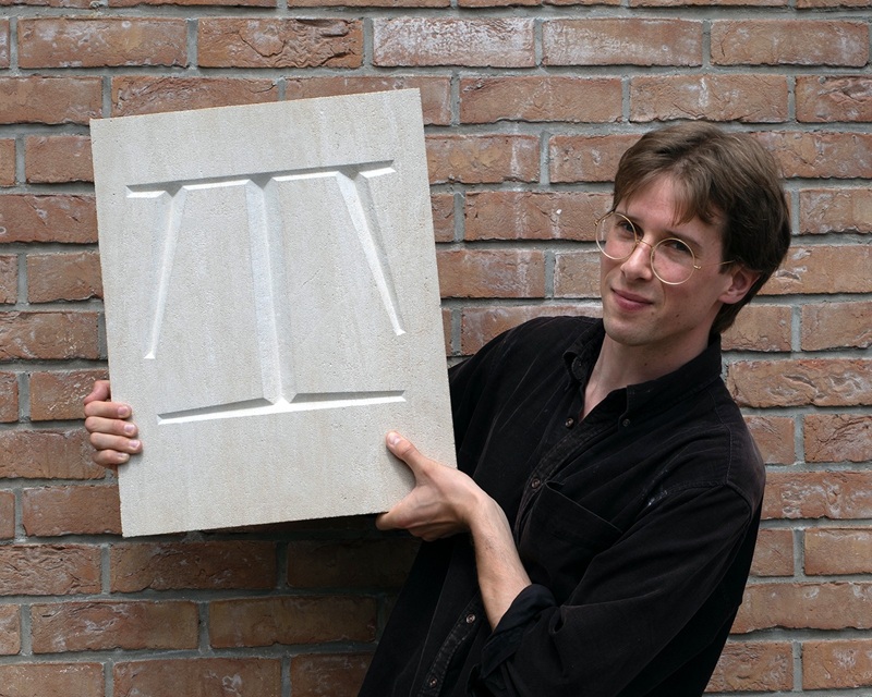 Person with short hair and glasses holds up plaque of an ornate letter "T". 