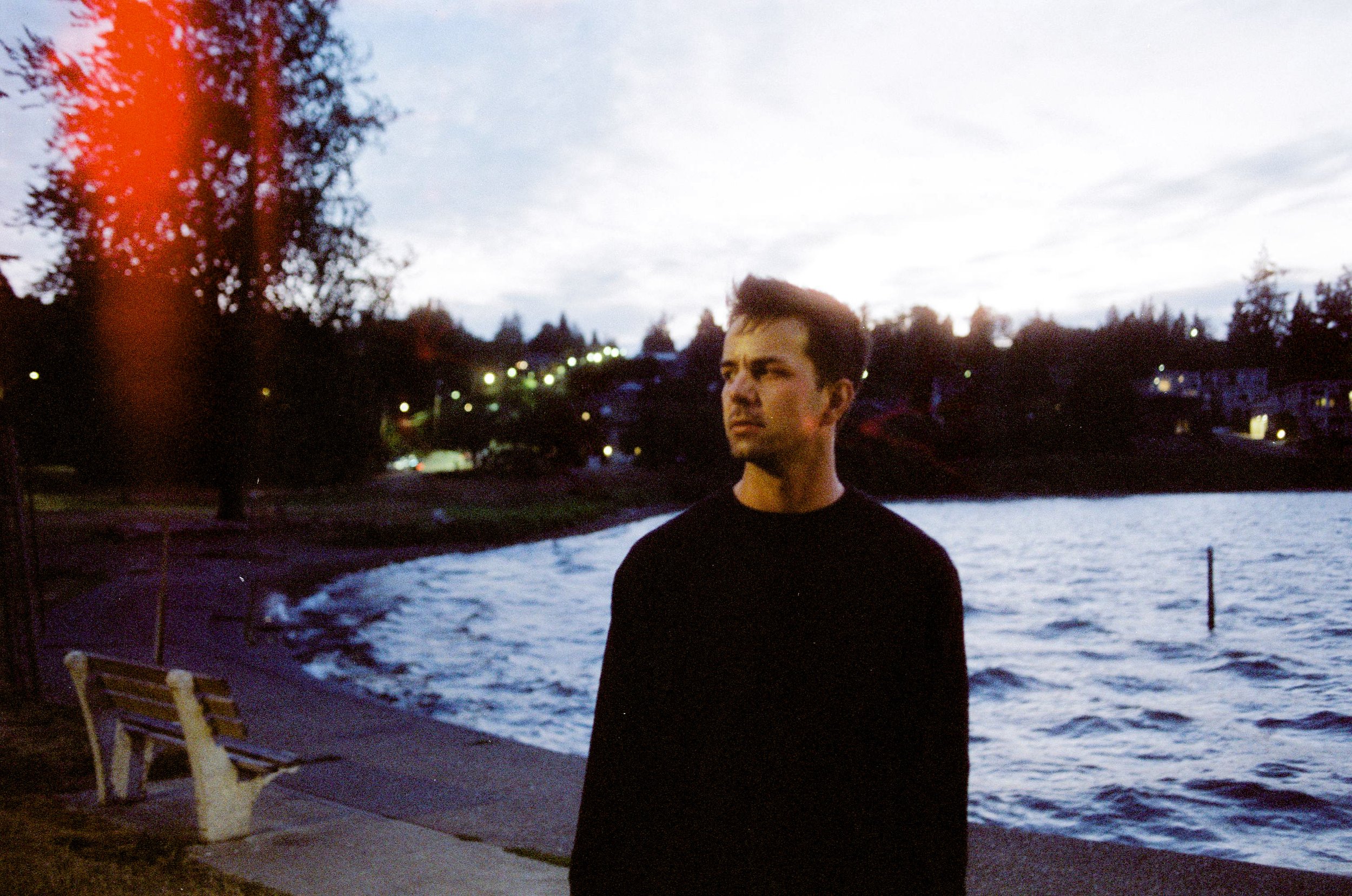 Young man with short hair and fair skin looks to side with lake in background, low light.  