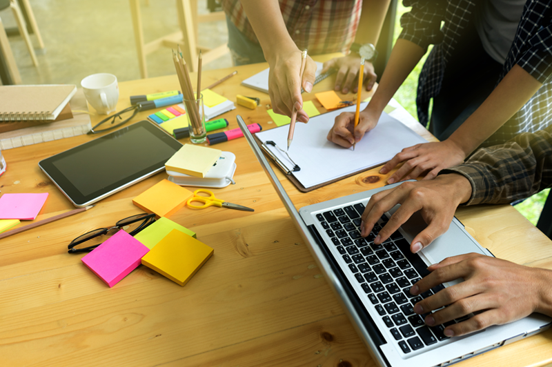 Three people brainstorming at a desk with a laptop, notepad, tablet, and colorful office supplies.
