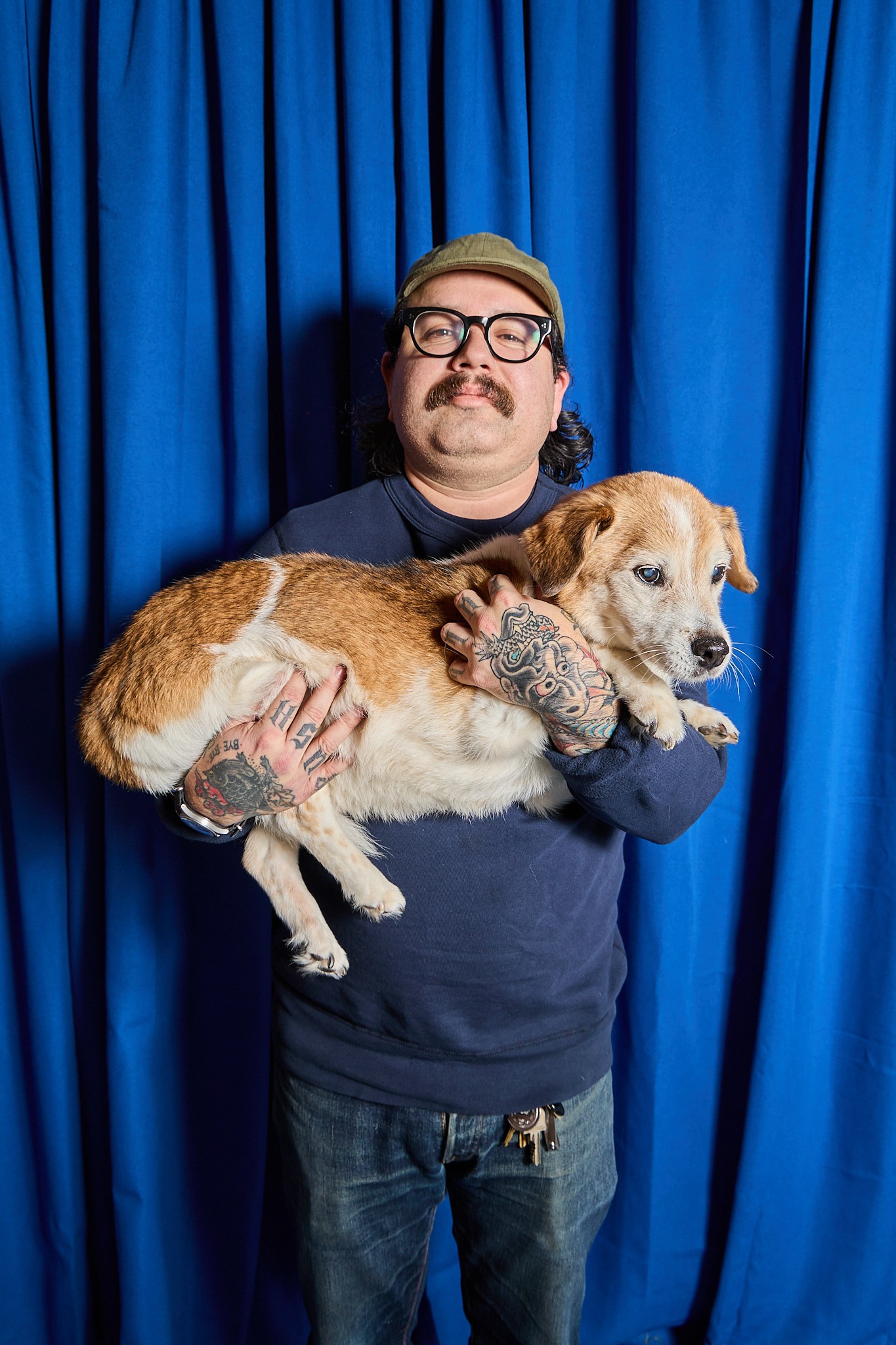 Photo of man with light skin, glasses and moustache holding a dog, showing tatooed hands, with blue drapes in the background. 