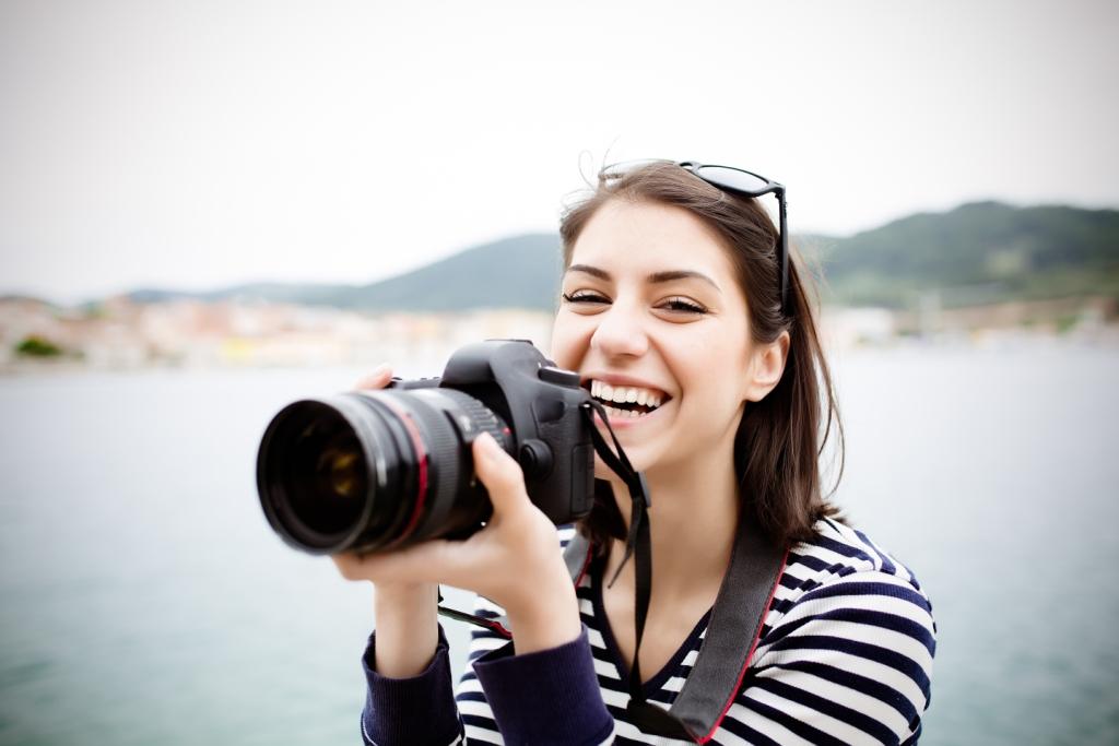 Smiling woman with light skin holds camera with large lens with lake and hills in the background.