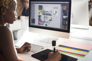 Woman with dark skin works at a desktop computer with images on the screen.  