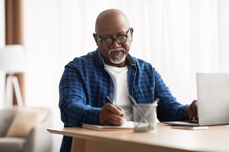 Man with dark skin writes on notepad with other hand on laptop computer. 