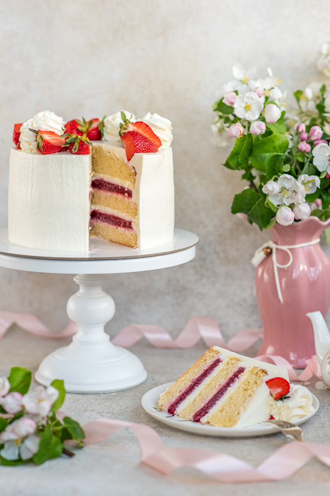 White cake on pedestal with slice on plate, red and white layers inside and strawberries and frosting flowers on top. 