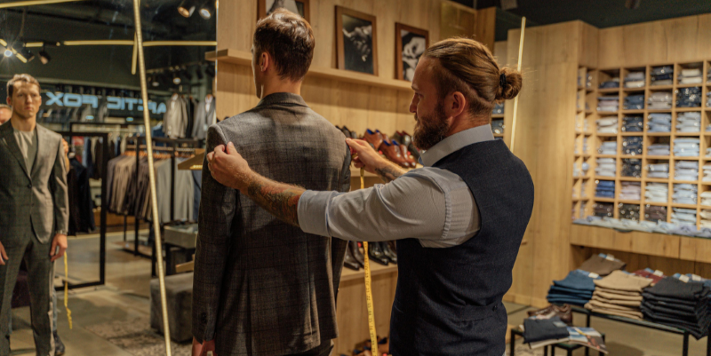 A man measures the shoulders of a man in a suit looking in the mirror.