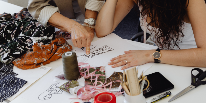 Two people at sewing table point to drawing with fabric and supplies around it. 