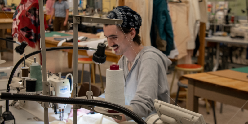 Student smiles while sewing at industrial machine in sewing classroom at Seattle Central College's Apparel Design & Development program. 