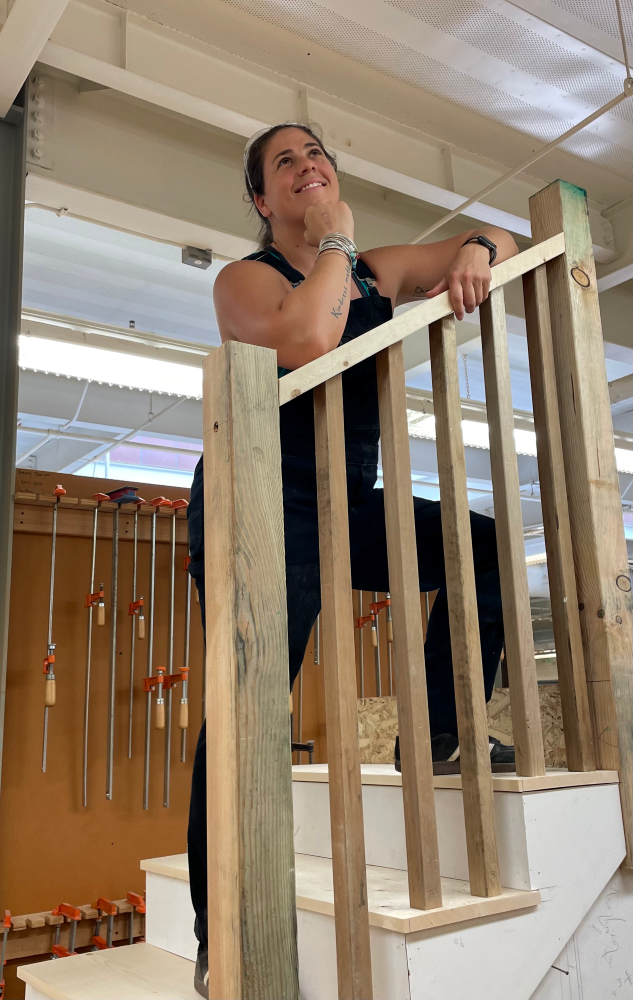 Person with light skin color stands on staircase looking upwards with a smile, with head in hand and arms resting on wooden railing. 