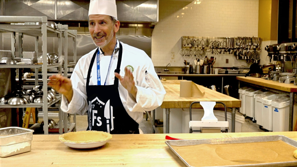 Man in professional kitchen wearing chef's hat and jacket and black apron smiles with a pie crust on the wooden table. 