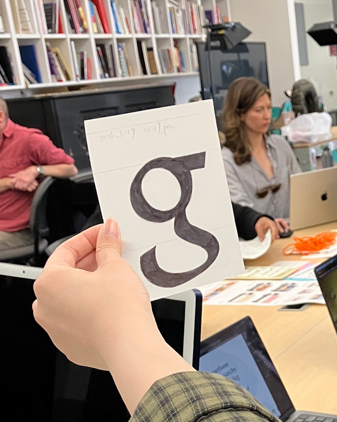 Someone with light skin holds a calligraphy "g" on a piece of paper with students sitting at tables and books in the background.  