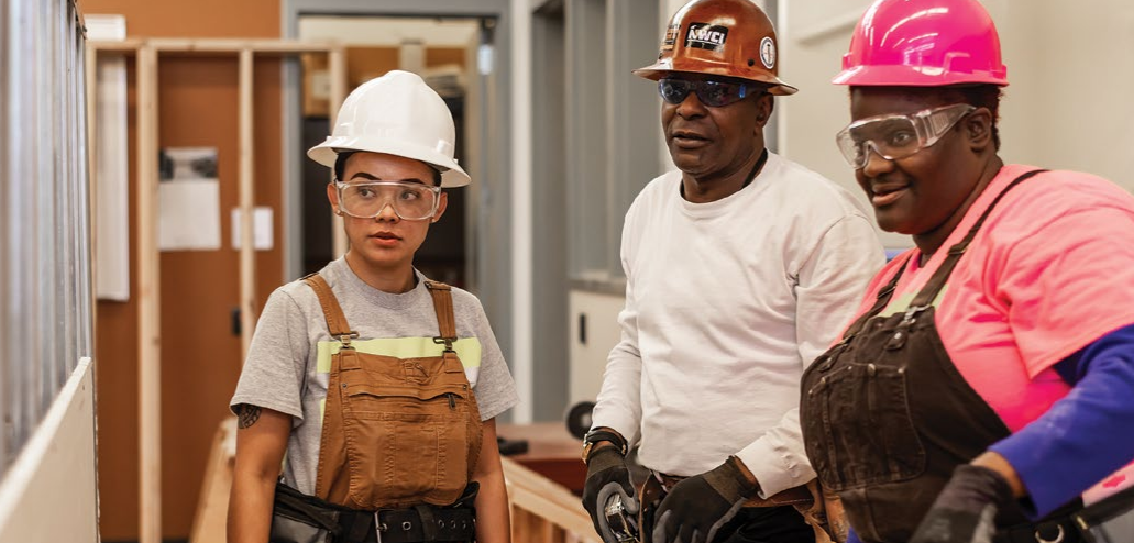 Three construction workers standing together with hard hats with wood framing behind them indoors.