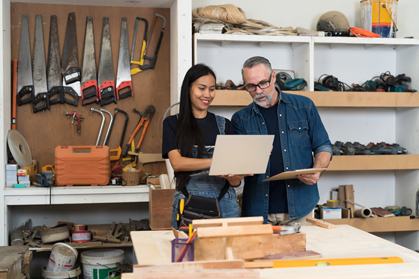 Man and woman in woodworking workshop standing at work table looking at laptop computer together. 